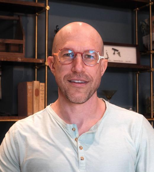 Headshot of Dr. O in a light green henley shirt wearing steampunk-esque orange, red, and gold glasses in front of a industrial chic bookshelf
