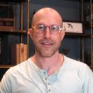 Headshot of Dr. O in a light green henley shirt wearing steampunk-esque orange, red, and gold glasses in front of a industrial chic bookshelf