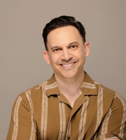 Smiling man in tan patterned shirt, arms crossed, beige studio headshot