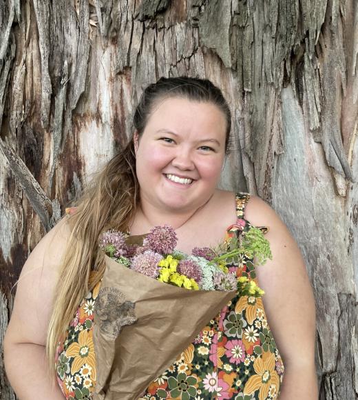 Erika, a fat white woman with brown hair and hazel eyes smiles brightly with the trunk of a large eucalyptus tree behind her. She is wearing orange, pink, and green floral dungarees and holding a pink, white, and yellow floral bouquet wrapped in brown paper. Her hair is tied in a low messy ponytail that drapes over her right shoulder.  