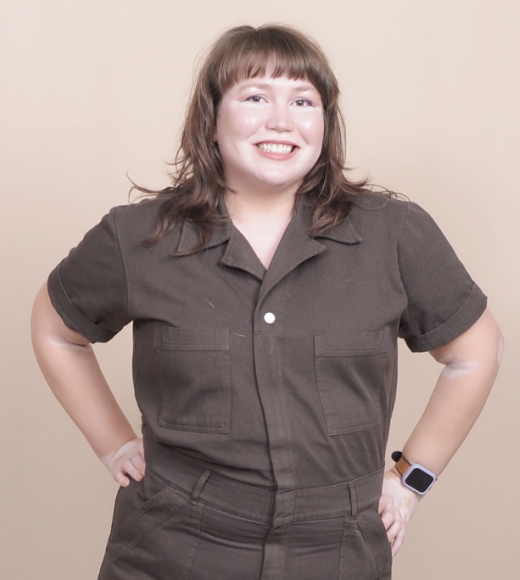 Elizabeth is seen from the waist up in an espresso colored cotton canvas short sleeve work jumpsuit. They pose with their hands on their hips. They have a layered light auburn brown fringe hair cut just below shoulder length.