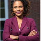 Headshot of Orly Clerge smiling at camera in a maroon blazer with her arms folded across her chest, with a white window frame and red brick wall in the background.