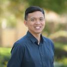 Headshot of Amir Aziz wearing a dark blue shirt with blurred trees and greenery in the background.