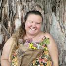 Erika, a fat white woman with brown hair and hazel eyes smiles brightly with the trunk of a large eucalyptus tree behind her. She is wearing orange, pink, and green floral dungarees and holding a pink, white, and yellow floral bouquet wrapped in brown paper. Her hair is tied in a low messy ponytail that drapes over her right shoulder.  