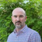 Man with a short beard wearing a checkered shirt, standing against a green leafy background.