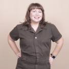 Elizabeth is seen from the waist up in an espresso colored cotton canvas short sleeve work jumpsuit. They pose with their hands on their hips. They have a layered light auburn brown fringe hair cut just below shoulder length.